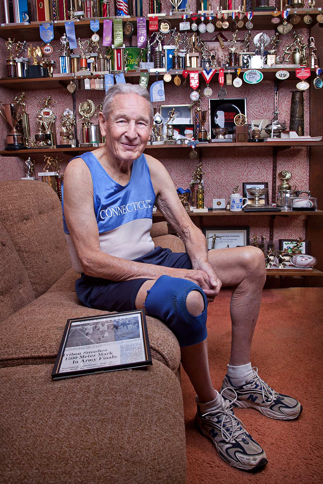 An accomplished elderly runner wearing his race outfit smiles with his trophies in his Connecticut home