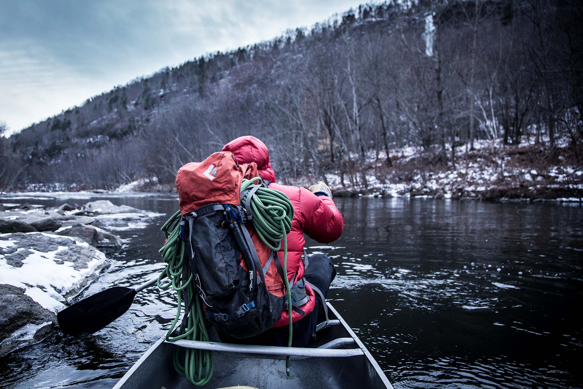 An ice climber in a canoe with climbing pack and rope paddles a river in winter